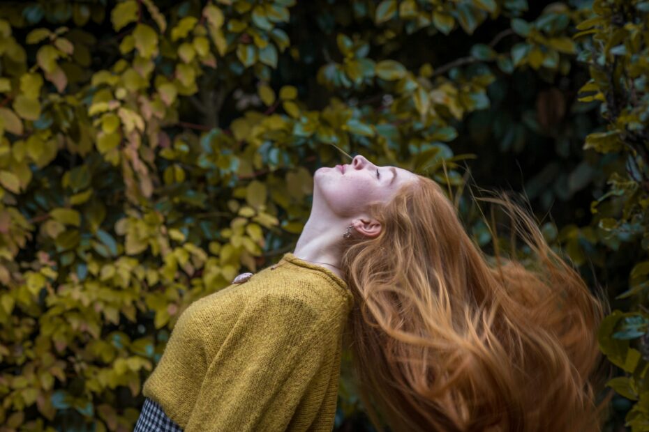 a red-haired woman wearing brown crew-neck shirt standing in front of green and yellow leaves